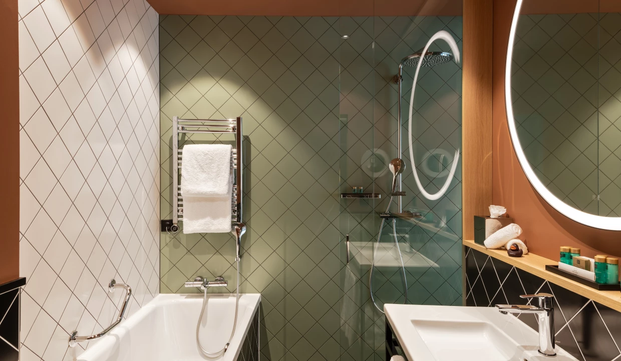 Hotelbad mit Wanne, diagonalen Fliesen in grün, salbei und terracotta. Beleuchteter runder Spiegel.