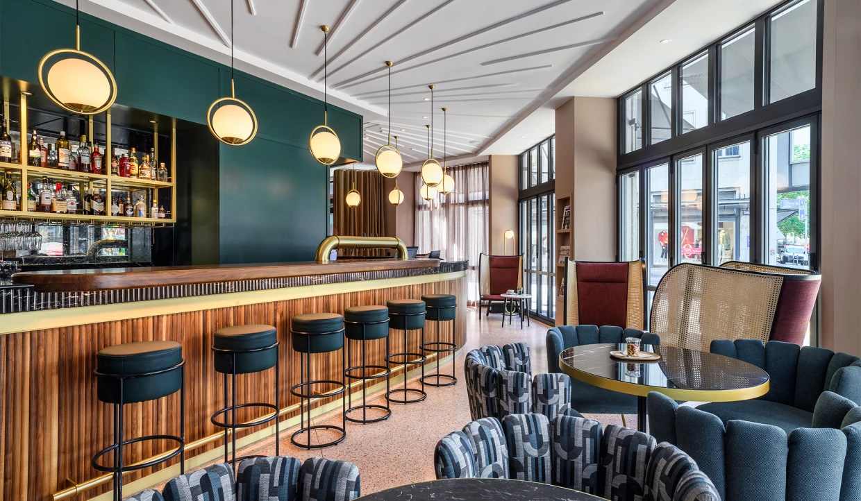 Langer Tresen einer Hotelbar mit Holzverkleidung mit Barhockern und Hängelampen. Davor Loungesessel im Art-Deco-Stil.