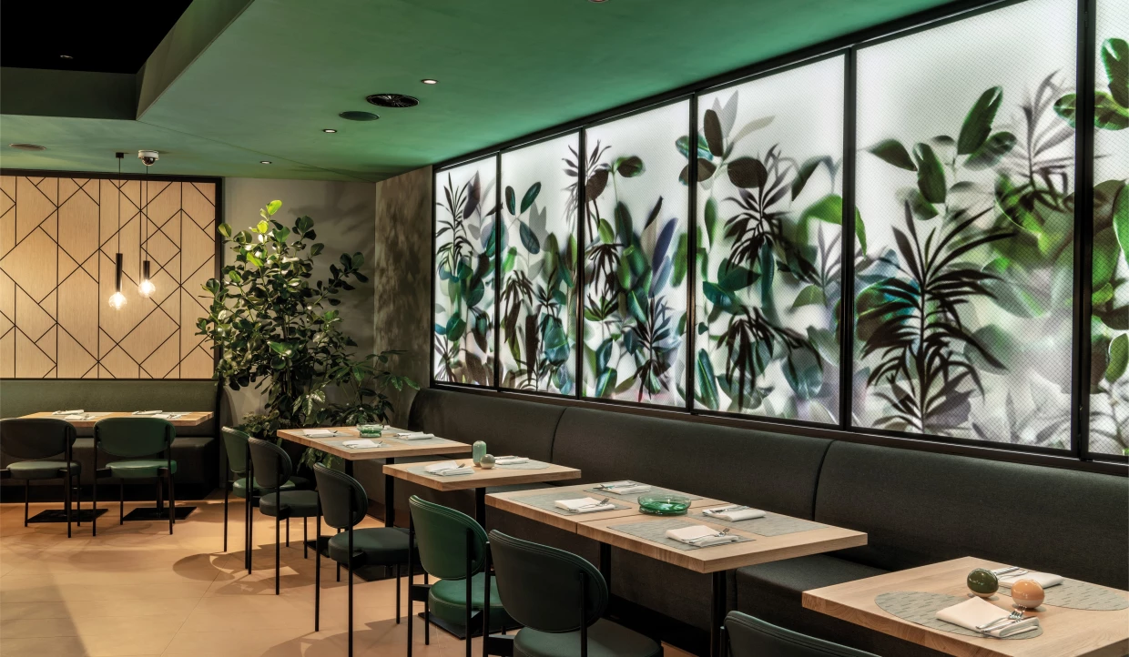 Modernes Hotel-Bistro mit Pflanzen, grünen Stühlen und einer Polsterbank und hinterleuchter Wand mit Pflanzenmotiven