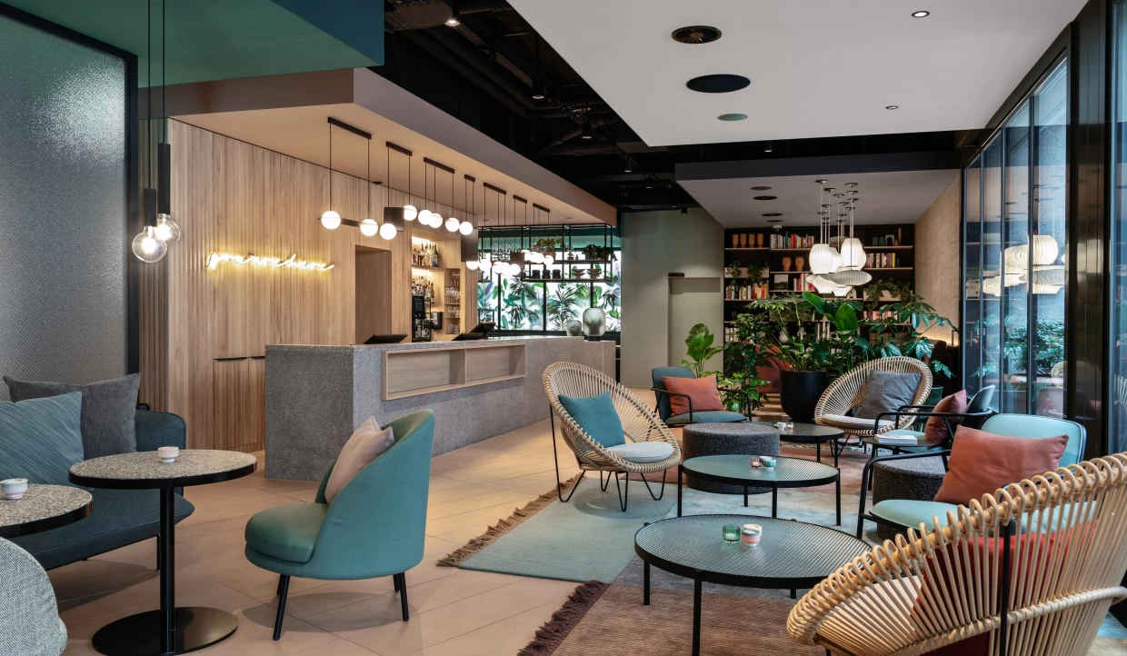 Reception eines modernen Stadthotels mit Holzvertäfelung, Korbsesseln und weiteren Loungemöbeln in türkis, beige und grau. 