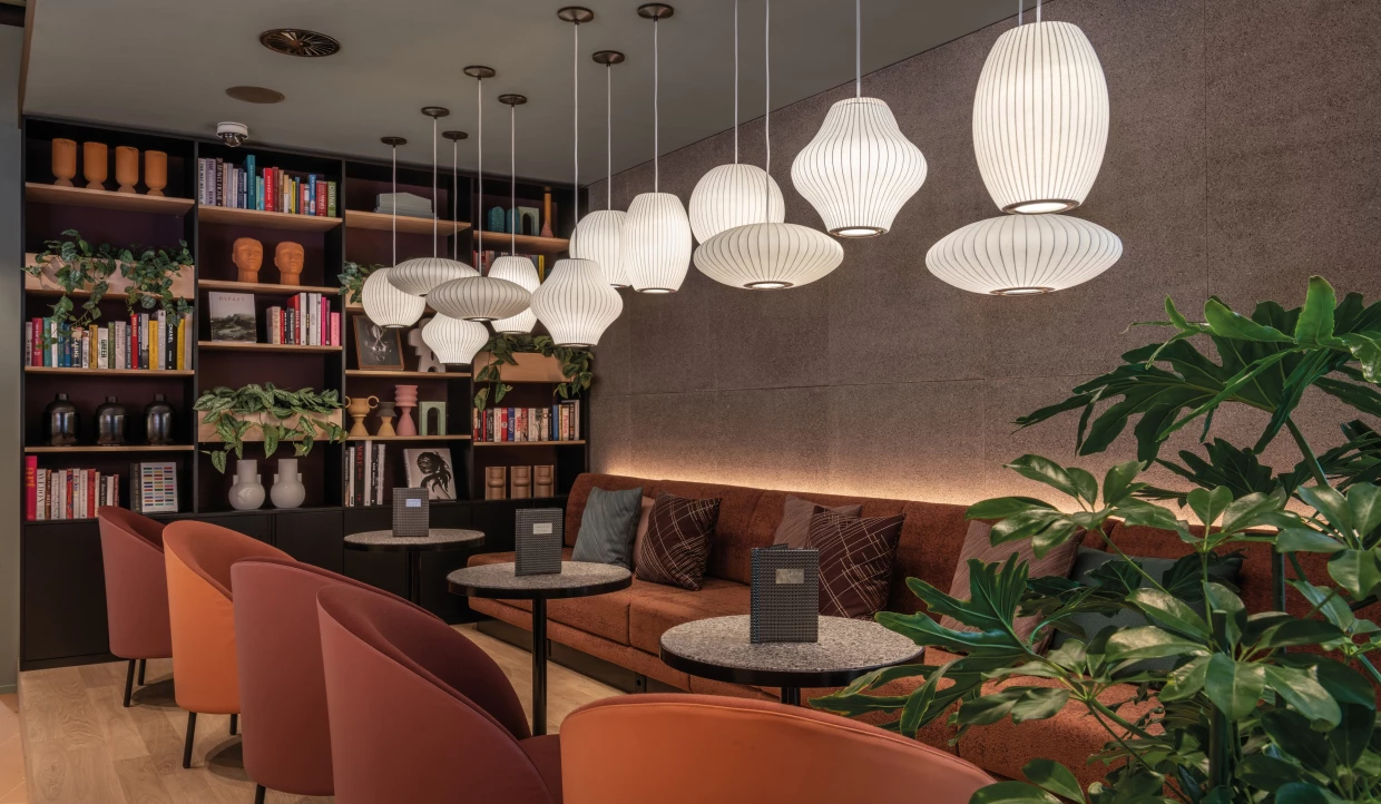 Stadthotel-Lobby mit weissen Hängelampen, terracottafarbenen Loungemöbeln, Bücherregalen und Pflanzen.