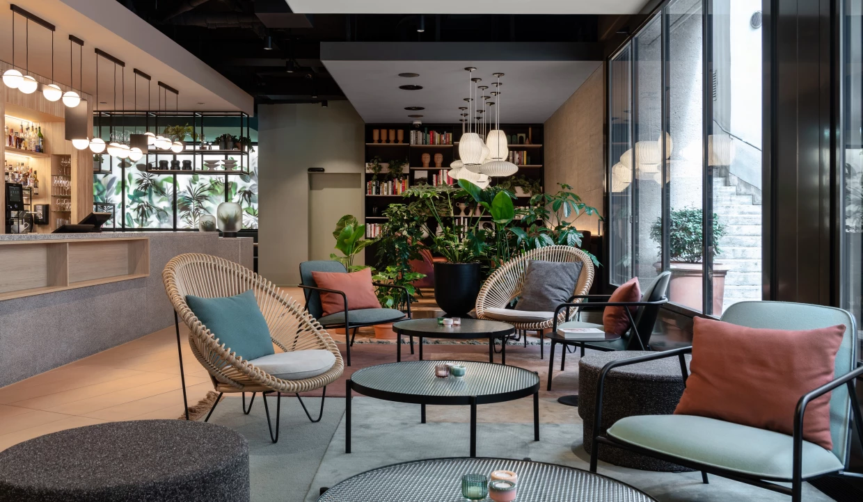 Moderne Lobby mit grossen Pflanzen, bodentiefen Fenstern, Loungesesseln und runden Tischen in terracotta, türkis und beige.
