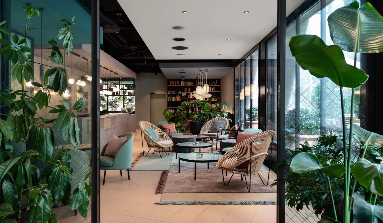 Hotel-Lobby mit Garten-Atmosphäre. Runde Tische, Pflanzen mit grossen Blättern und Rattansesseln.