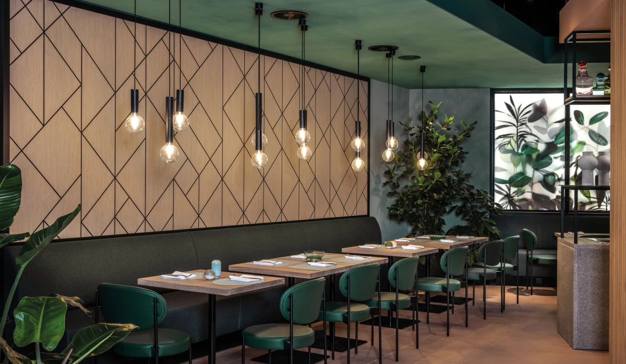 Modernes Bistro mit botanischem Flair, Holzvertäfelung mit Grafik, grünen Stühlen, Bank und Pflanzen.