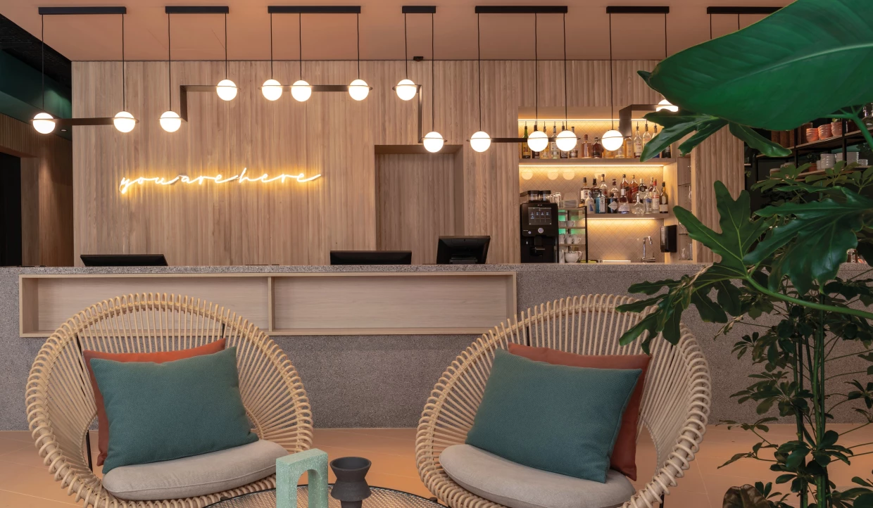 Reception eines Stadthotels mit Theke in Waschbeton, hölzener Verkleidung, davor Korb-Loungesesseln und Pflanzen