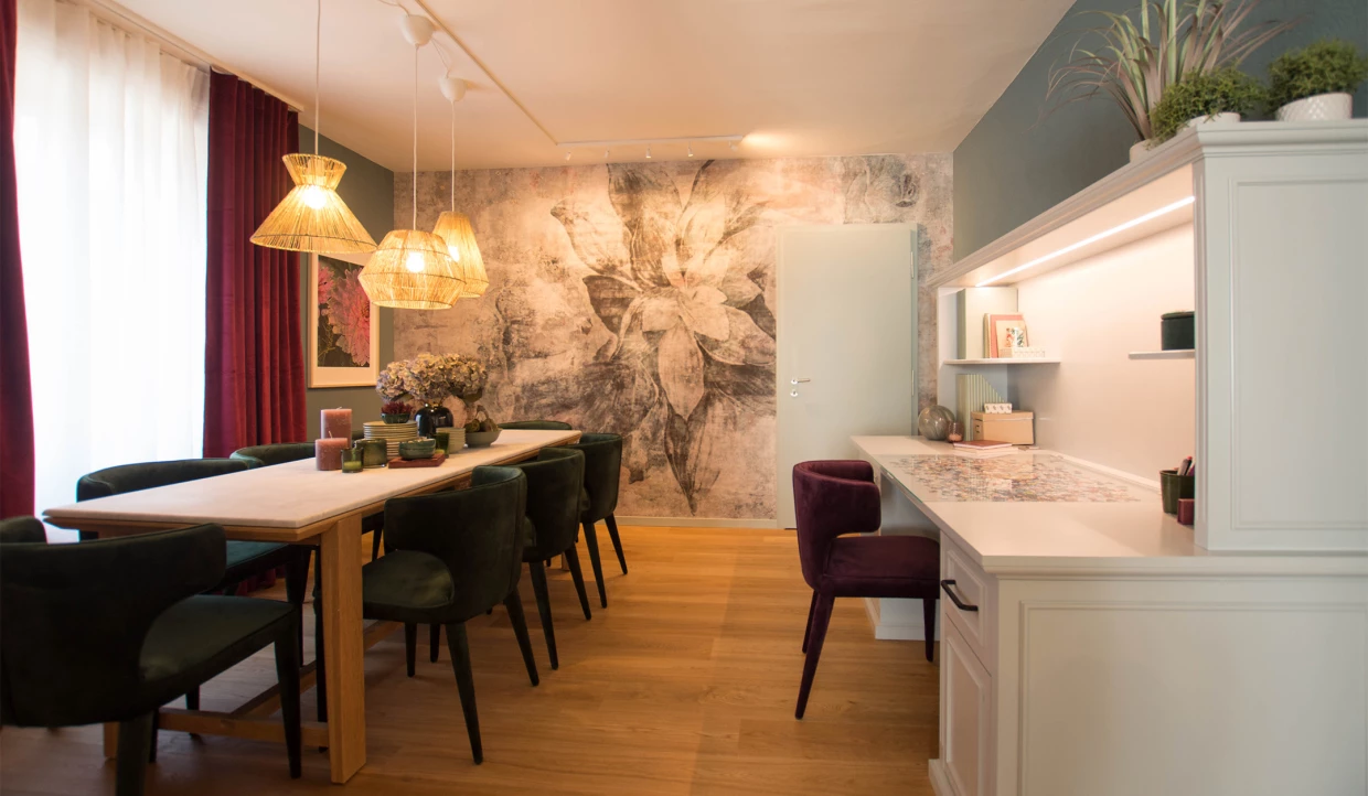 Esszimmer mit großem Holztisch, grünen Stühlen, hängenden Lampen und Wandbild