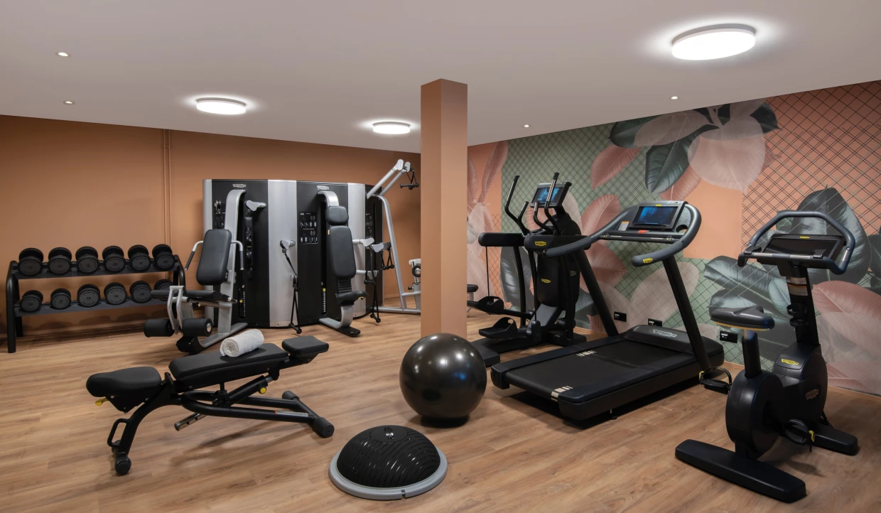 Hotel-Fitnessraum mit Geräten. Im Hintergrund eine Wand mit Pflanzenmotiven in apricot und grün. 