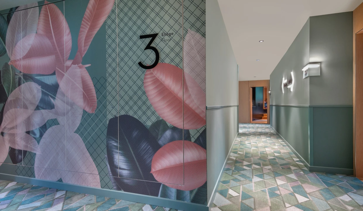 Blick in einen Hotelflur: Links eine Wand mit einer bunten Tapete mit grafischen und botanischen Motiven. Rechts ein langer Flur mit Teppich mit diagonalem Muster. 