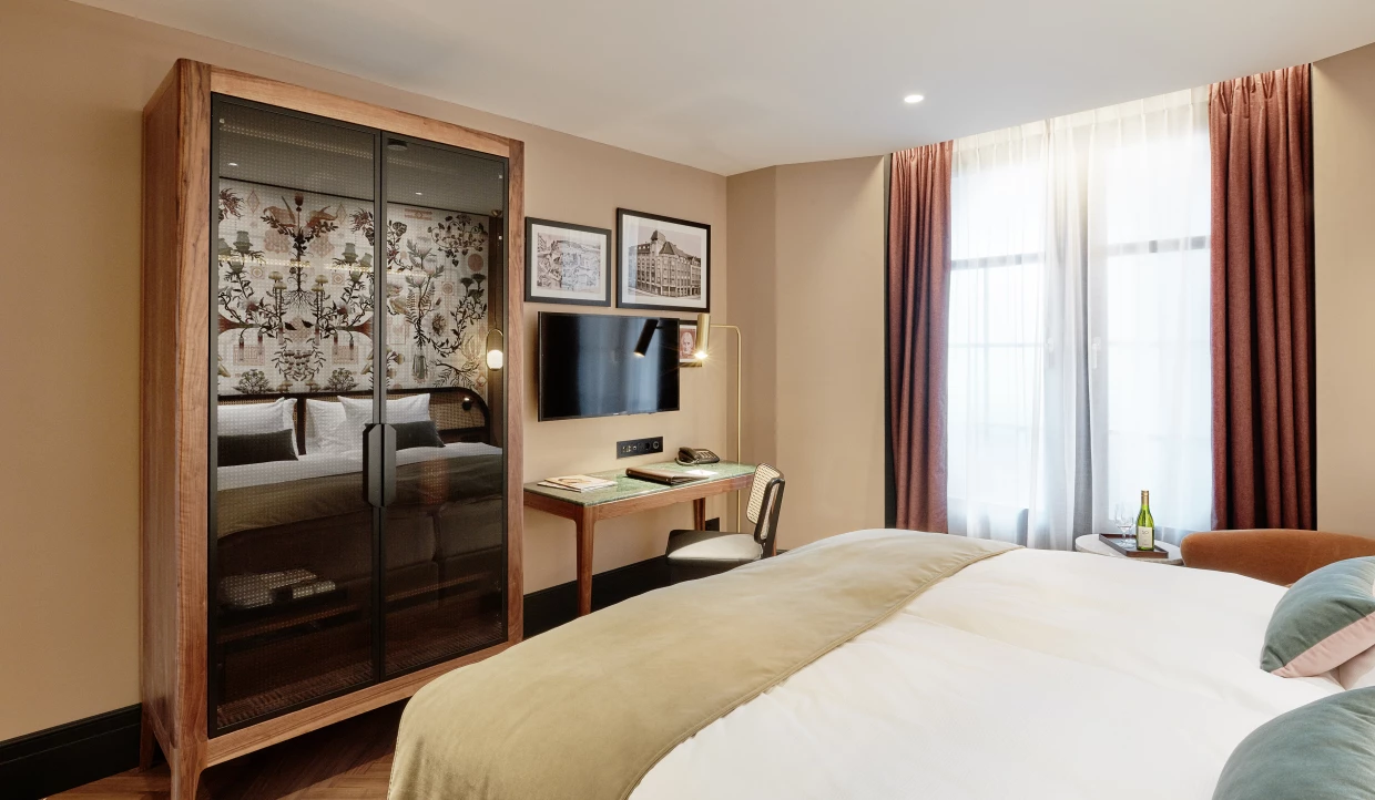 Hotelzimmer mit Bett, Schreibtisch und verspiegeltem Schrank in warmen Tönen. In der Spiegelung ist die Tapete zu sehen.