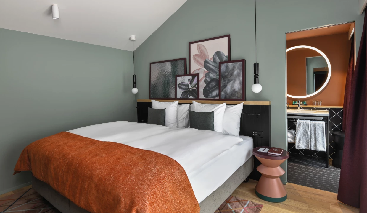 Modernes, frisches Hotelzimmer in orange, grau und türkis. Blick ins Bad. An der Wand Pflanzenmotive in Rahmen.