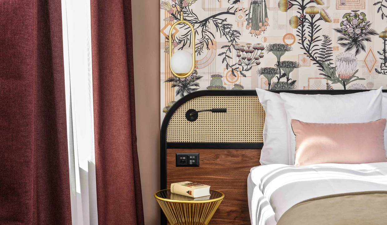 Kopfteil eines Hotelbetts in Bordeaux, Braun, Rosé und Beige mit Beistelltisch und Tapete in Art-Deco- und Jugendstilmustern.