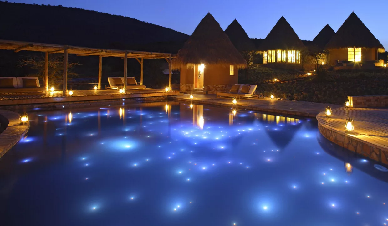 Pool eines Safari-Hotels mit beleuchteten Hütten in der Nacht.